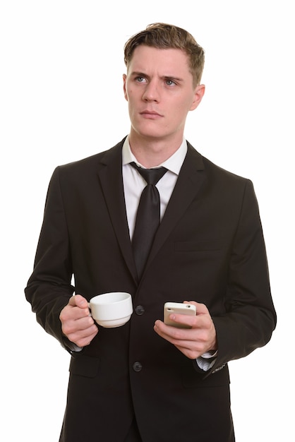 Joven empresario rubio guapo sosteniendo la taza de café mientras usa el teléfono