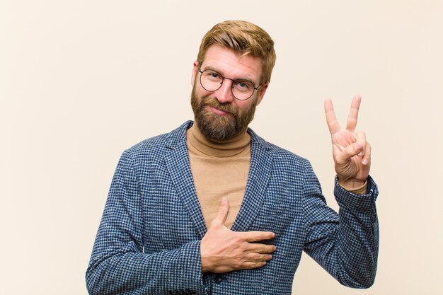 Joven empresario rubio que parece feliz, confiado y digno de confianza, sonriendo y mostrando el signo de la victoria, con una actitud positiva