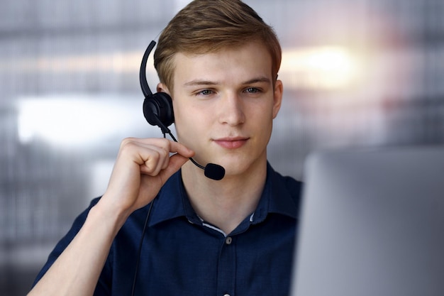 Joven empresario rubio usando auriculares y computadora en una oficina a oscuras, resplandor de luz en el fondo. Iniciar un negocio significa trabajar duro.