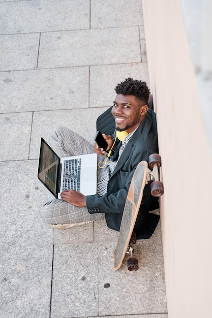 Joven empresario sentado al aire libre en una pared trabajando en una computadora portátil mirando a la cámara con un teléfono inteligente