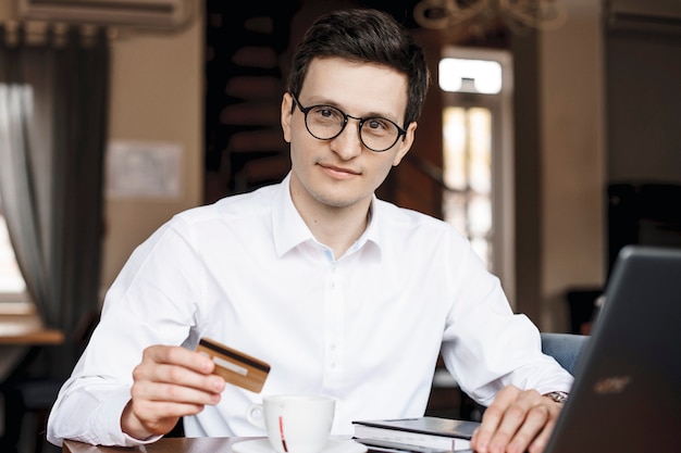 Foto joven empresario está sentado en una cafetería con una tarjeta de crédito en la mano y mirando a la cámara
