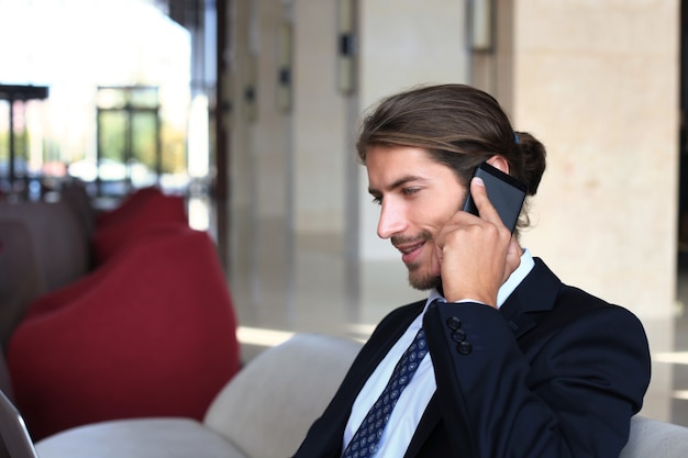 Joven empresario sentado relajado en el sofá en el vestíbulo del hotel haciendo una llamada telefónica, esperando a alguien.