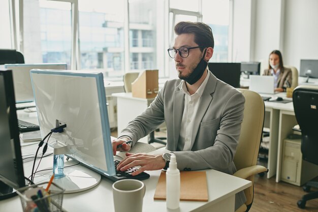 Joven empresario serio con máscara protectora negra debajo de la barbilla mirando la pantalla de la computadora mientras escribe en el lugar de trabajo en la oficina