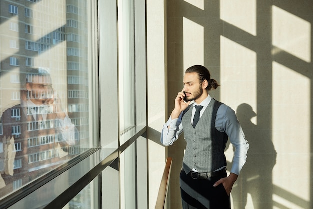 Joven empresario serio mirando por la ventana en un día soleado mientras consulta a uno de los clientes por teléfono inteligente en el día laborable