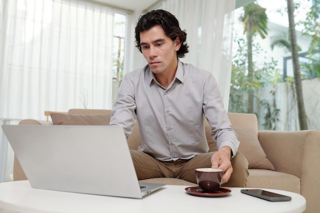 Joven empresario serio sentado en el sofá en el lugar de trabajo frente a la computadora portátil