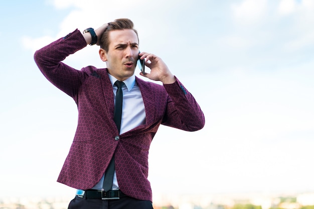 Foto joven empresario serio en traje rojo y camisa con corbata pararse en el techo y hablar por teléfono