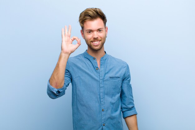 Joven empresario se siente feliz, relajado y satisfecho, mostrando aprobación con gesto bien, sonriendo contra la pared azul