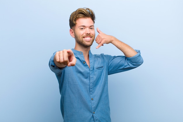 Joven empresario sonriendo alegremente y apuntando a la cámara mientras hace una llamada más tarde gesto, hablando por teléfono contra la pared azul