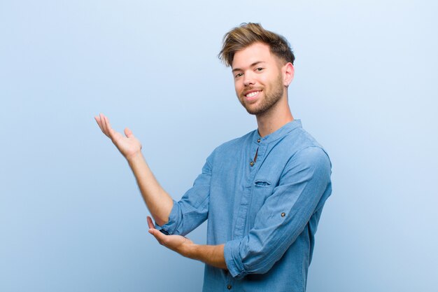 Joven empresario sonriendo con orgullo y confianza, sintiéndose feliz y satisfecho y mostrando un concepto en el espacio en blanco contra la pared azul