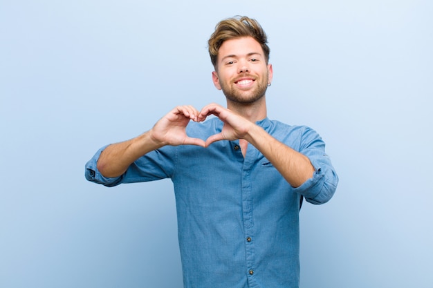 Joven empresario sonriendo y sintiéndose feliz, lindo, romántico y enamorado, haciendo forma de corazón con ambas manos contra la pared azul