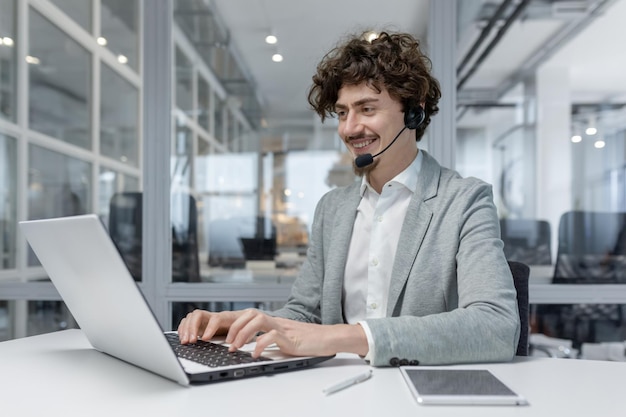 Un joven empresario sonriente con auriculares trabajando en una computadora portátil en una oficina moderna que muestra la multitarea