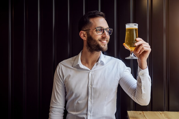 Joven empresario sonriente en camisa sentado en el pub después del trabajo, sosteniendo y mirando un vaso de cerveza y relajándose después del trabajo.