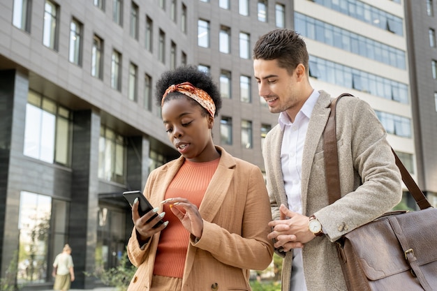 Joven empresario sonriente mirando la pantalla del teléfono inteligente en manos de su colega africano desplazándose a través de la información en línea