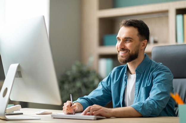 Joven empresario sonriente tomando notas mientras trabaja en la computadora en la oficina