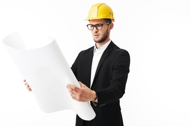 Joven empresario sorprendido en anteojos y casco de seguridad amarillo sosteniendo cuidadosamente el plan del nuevo proyecto en las manos