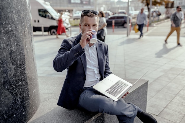 Joven empresario sosteniendo su computadora portátil mientras toma café. Trabajo a distancia en el exterior.