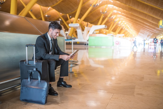 Joven empresario con tableta sonriendo en el aeropuerto y esperando su vuelo con equipaje