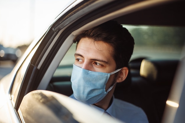 Joven empresario toma un taxi y mira por la ventanilla del coche con una máscara médica estéril. Concepto de distancia social.