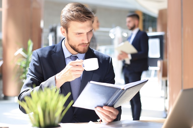 Joven empresario tomando café en la oficina mientras revisa el cuaderno.