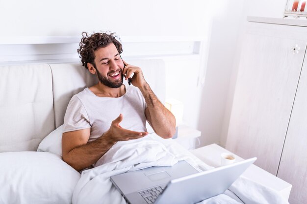 Joven empresario trabajando feliz y relajado desde la cama con ordenador portátil mediante teléfono móvil en concepto de adicto al trabajo, trabajando desde casa por la mañana y bebiendo café