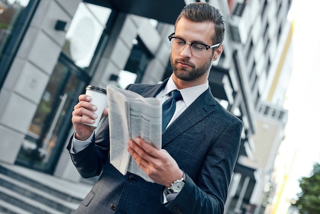 Joven empresario de traje y gafas sosteniendo una taza de papel y leyendo el periódico comercial en