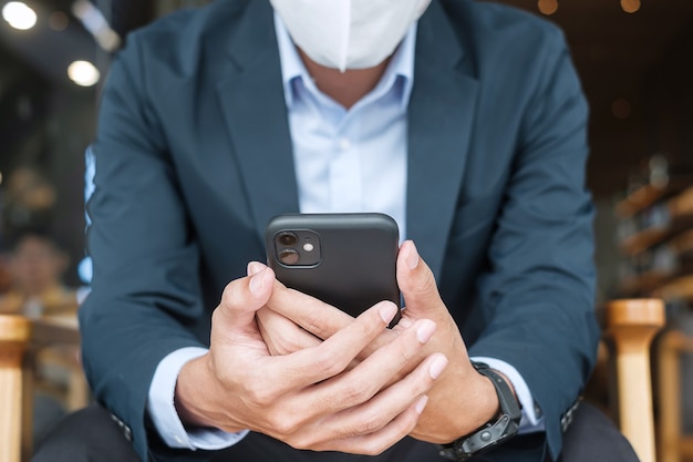 Joven empresario en traje con mascarilla quirúrgica y smartphone, hombre escribiendo teléfono móvil con pantalla táctil en la oficina o cafetería. Pandemia Covid-19, tecnología y nuevo concepto normal