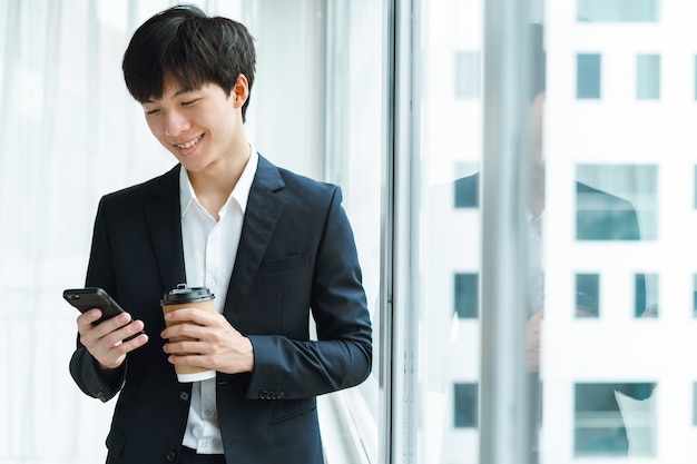 Joven empresario en traje tomando café y usando el teléfono junto a la ventana