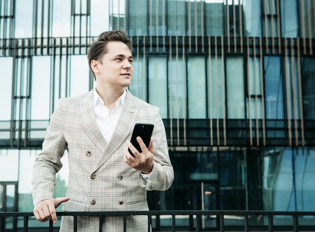 Joven empresario vestido con ropa formal parado afuera de un edificio de vidrio y usando teléfono móvil