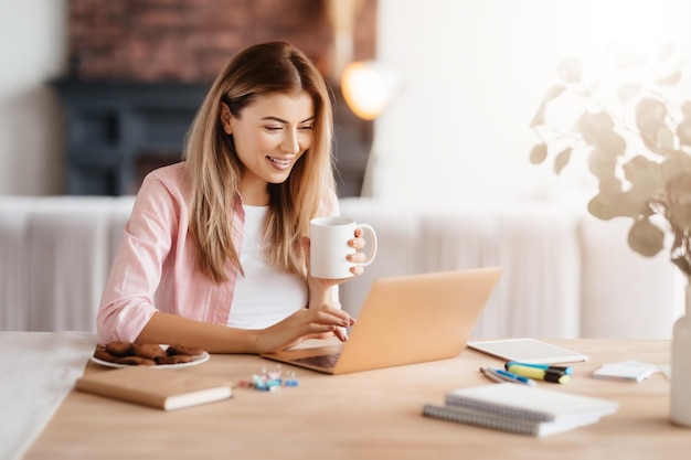 Una joven encantada con una taza de café sentada en el escritorio y mirando la pantalla del portátil