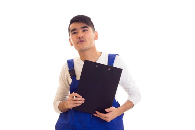 Un joven encantador con cabello oscuro vestido con camisa blanca y azul en general sosteniendo un bolígrafo blanco y una carpeta negra y hablando sobre fondo blanco en el estudio