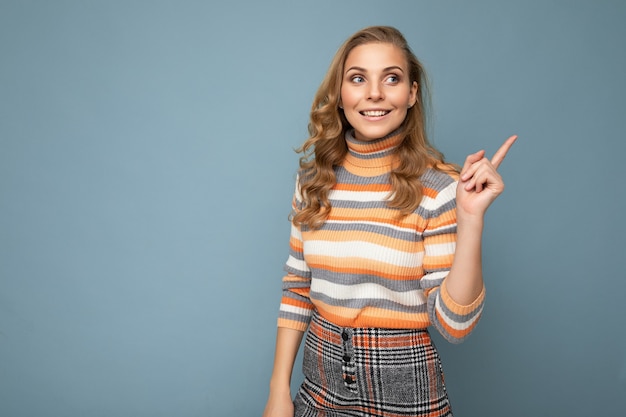 Joven encantadora fascinante sexy positiva feliz sonriente rubia mujer vestida con suéter de rayas casual aislado sobre fondo azul con espacio libre y señalar con el dedo en el espacio de la copia