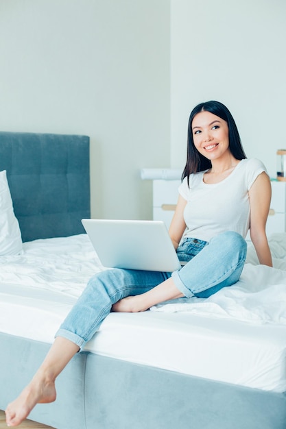 Una joven encantadora con ropa informal relajándose en la cama con una laptop en el regazo y mirando hacia otro lado con una sonrisa