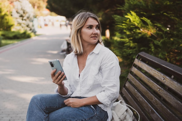 Una joven encantadora con ropa informal se sienta en un banco en el parque y usa un teléfono móvil para comunicarse