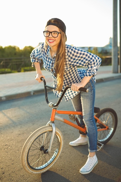 Foto una joven encantadora con un sombrero montando una bicicleta en el fondo de la ciudad al aire libre a la luz del sol