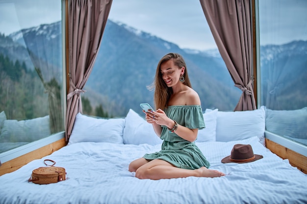 Joven encantadora sonriente hermosa boho chic morena mujer viajero con vestido esmeralda hombro desnudo sentado en la cama en la habitación del hotel con vista a la montaña y usando el teléfono para la comunicación en línea