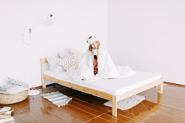 Una joven enferma con pijama rosa y una máscara para dormir en la cabeza está sentada en la cama