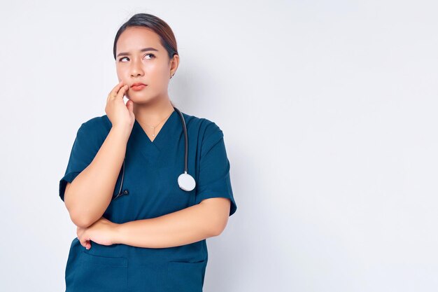 Una joven enfermera asiática molesta y molesta que usa uniforme azul con un estetoscopio se ve irritada o cansada, aburrida y exhala aislada en el fondo blanco Concepto de medicina de la salud