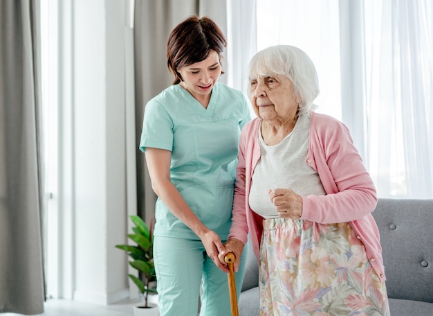 Joven enfermera ayuda a anciana en casa. La muchacha del trabajador de la salud se preocupa por la persona del sexo femenino mayor en el interior