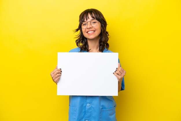 Joven enfermera doctora aislada de fondo amarillo sosteniendo un cartel vacío con expresión feliz