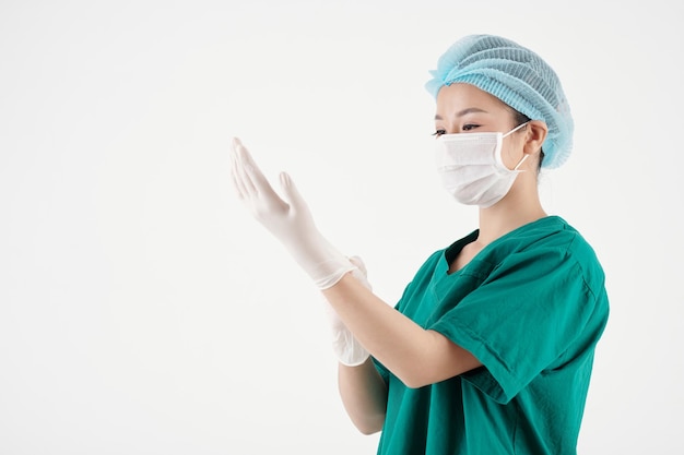 Joven enfermera con gorro médico y máscara protectora poniéndose guantes de goma al prepararse para el trabajo