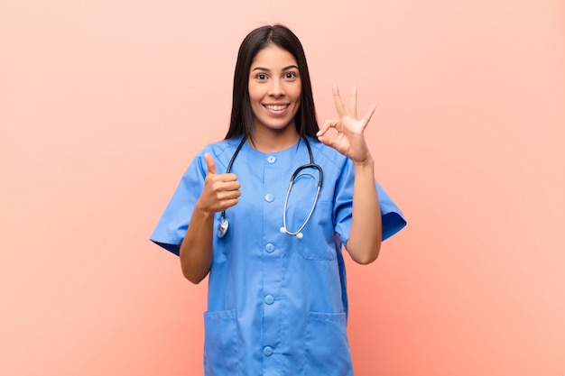 Joven enfermera latina se siente feliz, asombrada, satisfecha y sorprendida, mostrando gestos bien y pulgares arriba, sonriendo contra la pared rosa