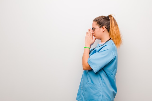 Joven enfermera latina sintiéndose preocupada, esperanzada y religiosa, rezando fielmente con las palmas presionadas, pidiendo perdón contra la pared blanca