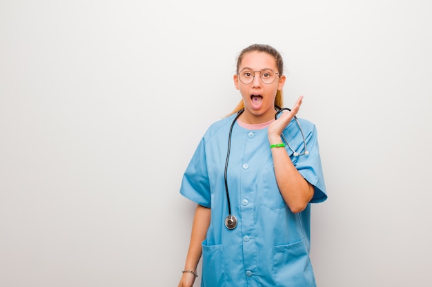 Joven enfermera latina sintiéndose sorprendida y emocionada, riendo, asombrada y feliz por una sorpresa inesperada en la pared blanca