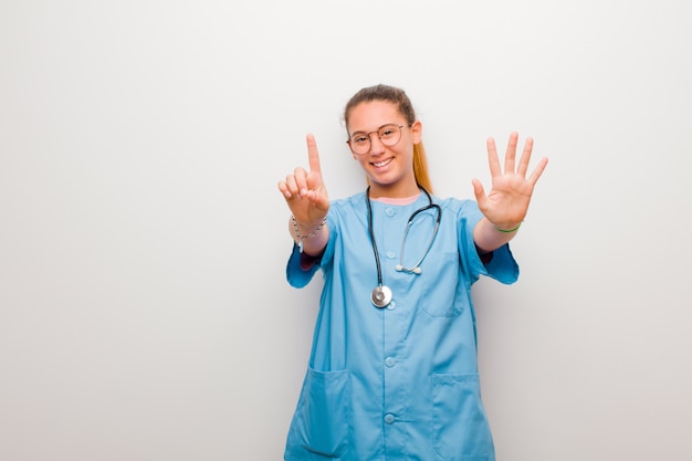 Joven enfermera latina sonriendo y mirando amigable, mostrando el número seis o sexto con la mano hacia adelante, contando hacia atrás en la pared blanca