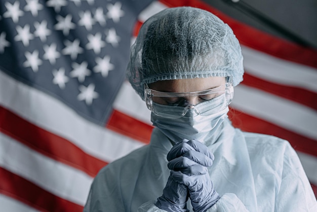 Joven enfermera con máscara médica protectora, sombrero y bata reza con la bandera estadounidense como telón de fondo y le pide a Dios ayuda y fuerza en la lucha contra la epidemia de coronavirus.