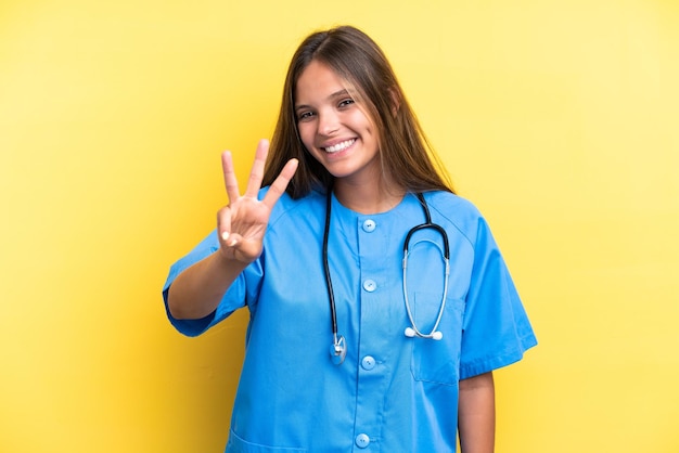 Joven enfermera mujer caucásica aislada de fondo amarillo feliz y contando tres con los dedos