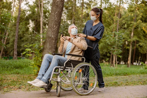 Joven enfermera en pantalla facial y mascarilla protectora ayudando a hombre discapacitado senior en silla de ruedas