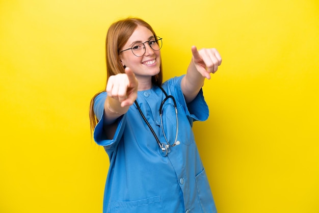 Una joven enfermera pelirroja aislada de fondo amarillo te señala con el dedo mientras sonríe