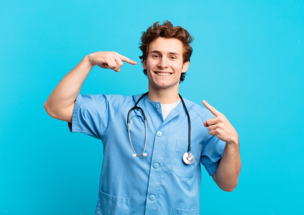 Joven enfermera sonriendo con confianza apuntando a su propia sonrisa amplia, actitud positiva, relajada y satisfecha