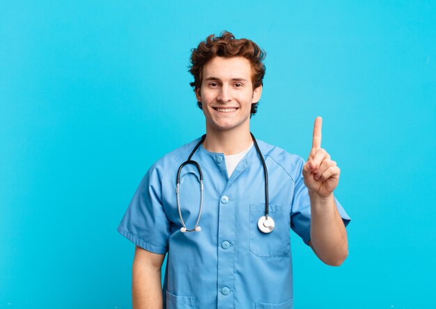 Joven enfermero sonriendo y mirando amigablemente, mostrando el número uno o el primero con la mano hacia adelante, contando hacia atrás